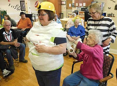 Myrtle Tebay, a resident of the Stewartville Care Center, seated in the foreground, wraps Rita Christian, Care Center activities assistant, in "mummy" toilet paper at a well-attended Halloween party at the Care Center on Thursday, Oct. 31. Tammy Wuerflin, activities assistant, stands behind Tebay. Care Center residents in the background, from left, beginning third from left, include Clem Snyder, Sandy Jones, Betty Fulk (partially hidden), and Betty Kraayenbrink.