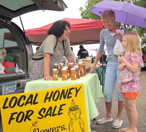 Sharon Peterson holds an umbrella to ward off the sprinkles as her granddaughter Arianna Woitas, 8, of Stewartville, contemplates a possible purchase from B's Honey, owned by Betty Kosnopfal of rural Chatfield, left. "We've built up quite a clientele," Kosnopfal said.