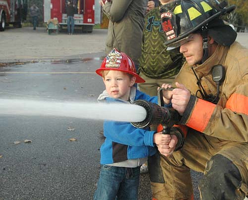 Jace Nagel, 3, of Stewartville, wearing the blue coat and firefighter hat, is one of hundreds of Stewartville and area children who practiced operating a fire hose with assistance from firefighter Nathan Ramaker at the Stewartville Fire Department's annual open house on Wednesday, Oct. 12. The event is held each year to celebrate Fire Prevention Week.