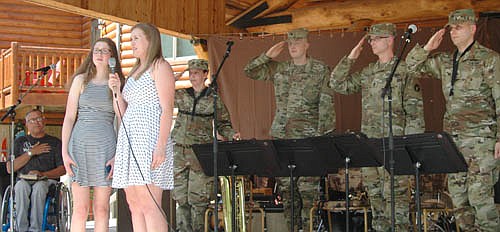 Emma Welch, a 2018 graduate of Stewartville High School, in the light-colored dress, and Megan Giordano, a junior at SHS, sing the National Anthem at the Ironwood Springs Christian Ranch Memorial Bash on Saturday afternoon, May 26. In the background are Bob Bardwell, founder of Ironwood Springs, far left, along with the saluting members of the Minnesota National Guard Band.